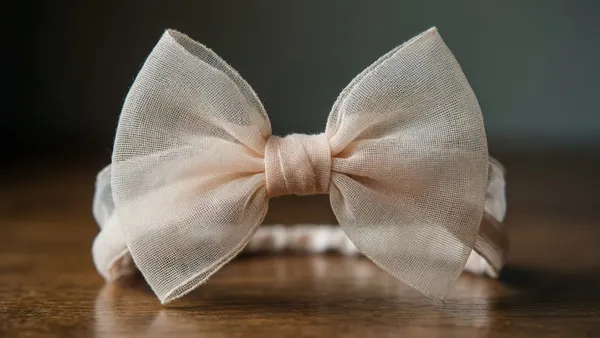 Laço de chiffon transparente em camadas dupla sobre faixa de tricoline, ao lado de tesoura de picotar e fita métrica em bancada de ateliê